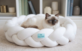 Вижте изображението в галерията, Cat lying on a braided white pet bed with a visible brand tag Furry Cuddle in a home setting.
